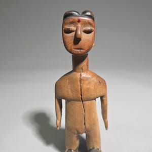 Könnte beinhalten: Eine handgeschnitzte Holzfigur mit hellbrauner Oberfläche, einem vertikalen Riss in der Mitte des Torsos, einem kleinen roten Punkt auf der Stirn, dunkel bemaltem Haar und dunkel bemalten Füßen, in aufrechter stehender Position.