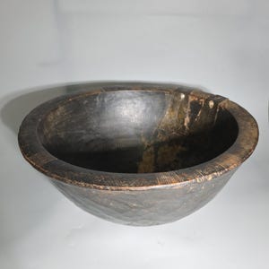 Puede incluir: Un cuenco grande de madera oscura con un borde ancho y un interior profundo. El cuenco tiene una superficie texturizada y veta de madera visible. El borde presenta líneas talladas. El cuenco es probablemente hecho a mano y tiene un aspecto rústico.