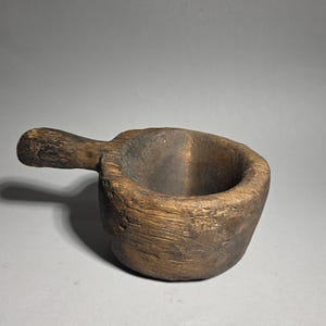 Può includere: Un piccolo mestolo in legno intagliato a mano di colore marrone scuro, con un manico corto e spesso e una coppa profonda e arrotondata. La superficie esterna presenta una consistenza ruvida e usurata con venature del legno ben visibili.
