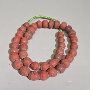 Könnte beinhalten: Eine Kette aus runden, verwitterten roten Perlen, die auf einer leuchtend lindgrünen Schnur aufgereiht sind. Die Perlen haben eine matte, strukturierte Oberfläche mit sichtbaren Alters- und Gebrauchsspuren, angeordnet in einer Kreisform.