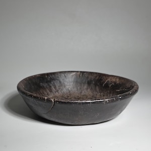 Könnte beinhalten: Eine dunkelbraune, rustikale Holzschale mit einem breiten, flachen Design. Die Schale hat eine glatte, polierte Außenseite und eine strukturierte Innenseite. Ein Riss ist an der Seite der Schale sichtbar. Die Schale ist wahrscheinlich handgefertigt und eignet sich zum Servieren von Speisen oder als Dekorationsstück.