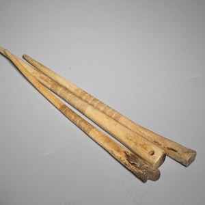 Könnte beinhalten: Drei antike, handgeschnitzte Holzwerkzeuge in hellbrauner Farbe. Die Werkzeuge haben eine lange, schlanke Form, eines davon mit einem Loch am Ende. Das Holz weist Alters- und Abnutzungserscheinungen auf, wobei einige Bereiche dunkler erscheinen.