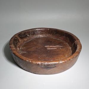 Könnte beinhalten: Eine rustikale, runde Holzschale mit dunkelbraunem Finish. Die Schale hat eine geringe Tiefe und einen breiten Rand, der die natürliche Holzmaserung und -struktur zeigt. Die Schale scheint handgeschnitzt zu sein, mit sichtbaren Unvollkommenheiten.