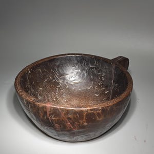 Könnte beinhalten: Eine handgeschnitzte Holzschale mit Griff. Die Schale hat eine dunkelbraune Farbe mit sichtbarer Holzmaserung und einer strukturierten Oberfläche. Die Schale hat ein rustikales, antikes Aussehen.