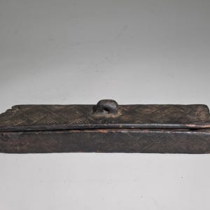 Könnte beinhalten: Eine rechteckige, dunkelbraune Holzschatulle mit einem Deckel, der einen mittigen Griff aufweist. Die Außenflächen von Deckel und Boden sind mit komplexen, geschnitzten geometrischen Mustern verziert und zeigen eine verwitterte Textur.