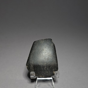 May include: A dark grey, irregularly shaped stone object, possibly a carving or artefact, is displayed on a clear acrylic stand. The stone has a textured surface and a slightly glossy appearance, set against a neutral grey backdrop.