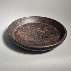 以下が含まれることがあります： 深さの浅い、濃い茶色の丸い木製のボウル。ボウルの内側は、木目が見え、表面が少しざらついた、質感のある風合いです。縁は滑らかで丸みを帯びています。