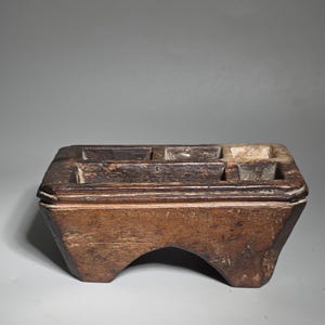 Könnte beinhalten: Eine verwitterte, rechteckige Holzkiste mit mehreren Fächern. Die Kiste hat eine dunkelbraune Farbe mit sichtbarer Holzmaserung und einen gebogenen Boden. Die Oberseite weist mehrere rechteckige Öffnungen auf, möglicherweise zur Aufbewahrung.