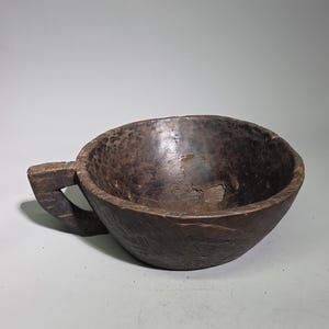 Könnte beinhalten: Eine dunkelbraune, handgeschnitzte Holzschale mit einem einzelnen, dicken und kantigen Griff an einer Seite. Die Schale weist eine raue, texturierte Außenseite mit sichtbaren Werkzeugspuren und ein tiefes, abgerundetes Becken auf.