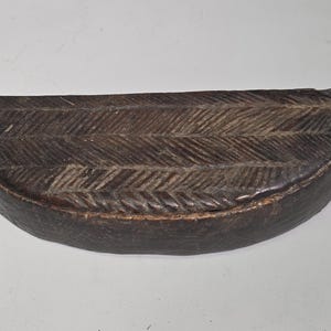 Könnte beinhalten: Eine halbkreisförmige, dunkelbraune Holzschatulle mit flacher Oberseite und gewölbtem Boden. Der Deckel ist mit einem geschnitzten Fischgrätenmuster über die gesamte Fläche versehen, während die Seiten der Schatulle glatt und unverziert sind.