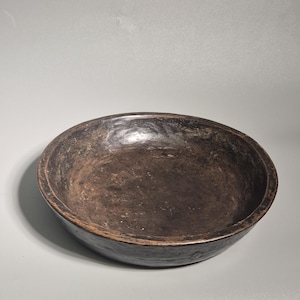 Può includere: Una ciotola rotonda in legno rustico con finitura marrone scuro. La ciotola ha un design ampio e poco profondo e un interno liscio. L'esterno mostra segni di usura, suggerendo età e utilizzo. Questa ciotola artigianale è adatta per servire o esporre.