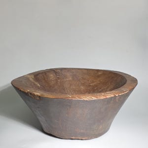 Könnte beinhalten: Eine große, antike Holzschale mit dunkelbrauner Außenseite und hellbrauner Innenseite. Die Schale zeigt die natürliche Holzmaserung. Die Form der Schale ist leicht konisch, mit einem breiten Rand.