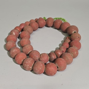 Op de afbeelding: Een streng van ronde, matte, terracotta-kleurige kralen met een ruwe, poreuze textuur. De kralen zijn geregen aan een lichtgroen koord en vormen een cirkelvorm. Elke kraal heeft een centraal gat en vertoont natuurlijke variaties in het oppervlak.