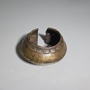 Op de afbeelding: Een brede, open messing armband met een verweerde, donkergouden patina. Het oppervlak is voorzien van een verhoogde, horizontale band versierd met een herhalend halfcirkelvormig, waaiervormig patroon rondom de hele omtrek.