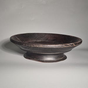 Könnte beinhalten: Eine dunkelbraune Holzschale auf einem Sockel mit einem breiten, flachen Becken. Die Schale wird von einem kurzen, abgerundeten Sockel getragen. Die Holzmaserung ist sichtbar und verleiht dem Stück Textur. Die Schale wird wahrscheinlich für dekorative oder servierende Zwecke verwendet.