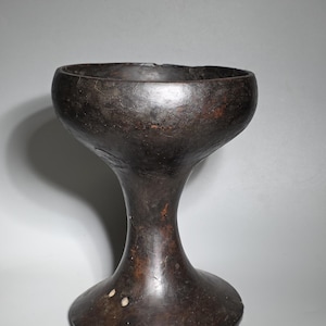 Può includere: Un calice artigianale marrone scuro con una coppa ampia e una base svasata. Il calice ha una superficie liscia e lucida e uno stelo che si allarga verso la base. La forma generale ricorda un calice o un vaso decorativo.
