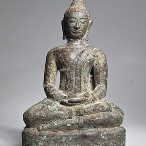 Könnte beinhalten: Bronze-Buddha-Statue in sitzender Meditationshaltung. Die Figur hat einen ruhigen Ausdruck, die Hände ruhen im Schoß. Die Statue hat ein verwittertes, gealtertes Aussehen mit Grün- und Brauntönen und eine spitze Kopfbedeckung.