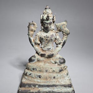 Könnte beinhalten: Eine verwitterte Bronzestatue einer sitzenden Gottheit mit mehreren Armen und einem detaillierten Kopfschmuck. Die Statue hat eine strukturierte Oberfläche mit grüner und dunkelbrauner Patina und ist auf einem quadratischen Sockel montiert. Die Figur befindet sich in meditativer Pose.