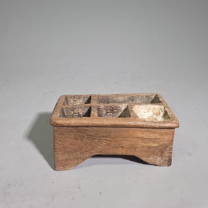 Può includere: Un organizer rettangolare in legno dotato di cinque scomparti distinti di varie dimensioni. Il legno marrone naturale presenta una superficie usurata e strutturata con venature visibili e una base robusta con un intaglio ad arco.