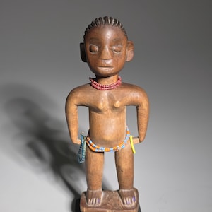 Könnte beinhalten: Eine geschnitzte Holzfigur einer Person mit geschlossenen Augen, die eine rote Halskette und Perlenverzierungen an Taille und Handgelenk trägt. Die Figur steht auf einem rechteckigen Sockel. Das Holz hat einen warmen Braunton.