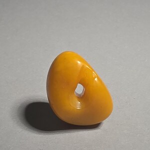 May include: A smooth, polished, golden-yellow amber-colored bead with an irregular, rounded, pebble-like shape. The object features a single, centered, circular hole that passes through the thickest part of the material, creating a hollow opening.