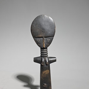 Könnte beinhalten: Eine dunkelbraune, geschnitzte Holzfigur mit einem großen, flachen, kreisförmigen Kopf, eingravierten dreieckigen Augen und einer vertikalen Nasenlinie. Der Hals ist verlängert mit horizontalen Ringdetails, die zu einem Torso mit kleinen Brüsten führen.