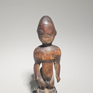 Könnte beinhalten: Eine geschnitzte Holzfigur mit rundem Kopf und stilisierten Gesichtszügen. Die Figur hat einen muskulösen Körperbau und steht auf einem rechteckigen Sockel. Das Holz hat einen warmen, braunen Farbton mit sichtbarer Maserung und Abnutzung. Die Figur steht aufrecht.