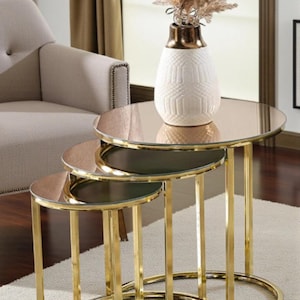Può includere: Set di tre tavolini a incastro con struttura in metallo dorato e piani rotondi in vetro a specchio. Un vaso bianco con un motivo geometrico e fiori secchi è posto sul tavolo più grande. Tavoli per l'arredamento del soggiorno.