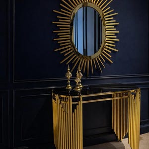 May include: A gold console table with a black top and an oval mirror with a sunburst design. Two decorative objects sit on the table. The wall is a dark blue colour. The table is likely made of metal.