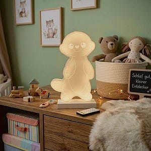 Könnte beinhalten: Ein leuchtendes Nachtlicht in Erdmännchenform auf weißer Basis, mit Fernbedienung. Das Licht steht auf einer Holzkommode mit gerahmter Tierkunst und einem Korb mit Stofftieren. Der Raum hat eine grüne Wand.