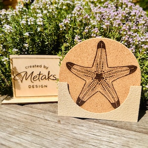 Könnte beinhalten: Ein runder Korkuntersetzer mit einem dunkelbraunen Seestern-Design, der in einem hellbeigen Halter liegt. Der Untersetzer und der Halter stehen auf einer Holzoberfläche, mit einem kleinen Holzschild mit der Aufschrift "created by Metaks DESIGN".