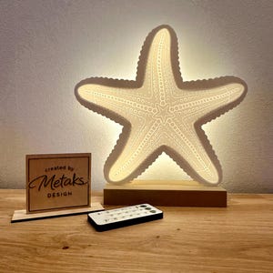 Può includere: Lampada a LED a forma di stella marina con intricati dettagli ritagliati, montata su una base di legno. Include un telecomando e un piccolo cartello di legno con la scritta "created by Metaks DESIGN".