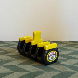 May include: A black and yellow ceramic toast rack with a Marmite jar design. The rack has four slots for toast and a yellow handle. The Marmite jar logo is visible on the side.