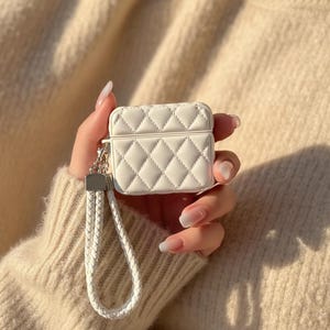 May include: A white, quilted AirPods case with a silver-tone clasp and a braided wrist strap. The case has a diamond pattern and a rectangular shape. The background is a soft, neutral color.