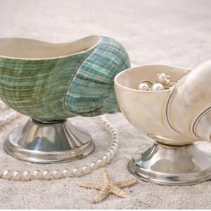 Puede incluir: Dos cuencos decorativos con forma de concha y bases plateadas. Uno es verde y el otro blanquecino, ambos llenos de joyas. Un collar de perlas y una estrella de mar están sobre la arena.