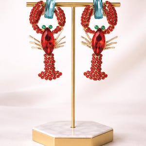 May include: A pair of lobster-shaped earrings. The earrings are red with gold accents and feature a large red gemstone at the centre. The lobster claws are adorned with red rhinestones, and the eyes are green. A light blue rectangular gemstone sits at the top.