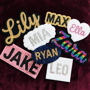 May include: A collection of assorted sequin patches featuring names including Lily, Max, Ella, Mia, Zara, Jake, Ryan, and Leo. The patches come in various shapes such as rectangular, oval, heart, and cloud, showcasing colors like gold, black, white, pink, and rainbow.
