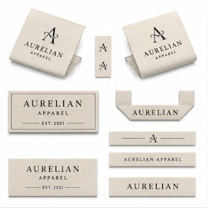 May include: A collection of beige fabric clothing labels featuring the black text AURELIAN APPAREL and the A logo. The set includes various rectangular, folded, and end-fold tags with different layouts, some displaying the text EST. 2021 with horizontal lines.