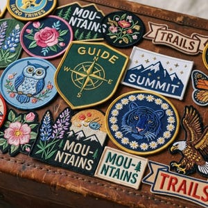 Op de afbeelding: Een verzameling geborduurde stoffen patches met diverse ontwerpen, waaronder een blauwe uil, een groen kompas met de tekst GUIDE, een tijger omringd door madeliefjes, een Amerikaanse zeearend en meerdere patches met de tekst MOUNTAINS, SUMMIT en TRAILS.