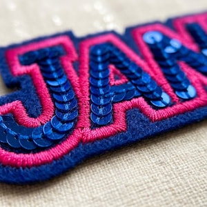 Könnte beinhalten: Ein Stoffaufnäher mit den Buchstaben J, A, N und E in einer fetten Schriftart. Die Buchstaben sind mit glänzenden blauen Pailletten gefüllt und mit leuchtend rosa Stickgarn umrandet, alles auf einer dunkelblauen Filzunterlage.