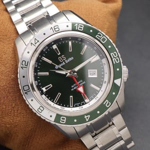 May include: A stainless steel Grand Seiko automatic watch with a green dial and a green and silver bezel. The watch has a date window at the 3 o'clock position and a silver bracelet. The watch is displayed on a brown surface.