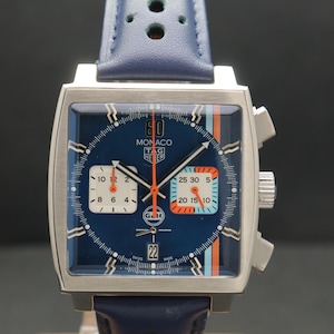 Op de afbeelding: Vierkante polshorloge met blauwe wijzerplaat en blauwe leren band. De wijzerplaat van het horloge heeft witte en oranje accenten, met de tekst "MONACO TAG HEUER". Het horloge heeft een zilverkleurige kast.