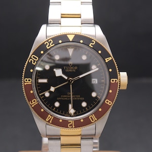 Puede incluir: Un reloj de pulsera bicolor de acero inoxidable y oro con esfera negra. El reloj presenta un bisel giratorio con marcas marrones y negras, detalles dorados y el texto "TUDOR GENEVE". La correa es una combinación de plata y oro.