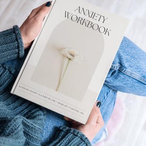May include: A person holds a cream-coloured "Anxiety Workbook". The cover displays the title in black text, a floral illustration, and the quote "Although anxiety is a part of life - never let it control you."