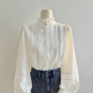 May include: A white blouse with long sleeves and a high, ruffled collar. The blouse features delicate lace detailing down the front and on the sleeves. It is buttoned up the front and is paired with blue jeans.