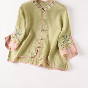 Op de afbeelding: Een lichtgroene linnen blouse met roze afwerking en bloemenborduurwerk. De blouse heeft een mandarijnkraag, kikkerknopen en driekwart mouwen met roze manchetten. De bloemenborduurwerk staat op de kraag en mouwen.