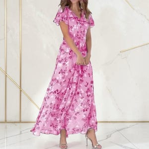 Op de afbeelding: Een lange, roze jurk met bloemenprint, fladderende mouwen en een V-hals. De jurk heeft een patroon van roze en witte bloemen. Het model draagt zilveren sandalen met hoge hakken.