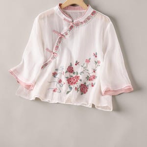May include: White embroidered blouse with a mandarin collar and three-quarter sleeves. The blouse has pink trim on the collar and sleeves, with floral embroidery in pink and green. It features a button closure and is made of delicate, flowing fabric.