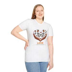 Può includere: T-shirt bianca con un disegno a forma di cuore di farfalle e mani. Il disegno include la parola "LOVE" e la frase "IS MORE THAN SKIN DEEP" in un carattere di scrittura. Le farfalle sono nelle tonalità dell'arancione, del marrone e del nero.