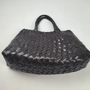 May include: A dark brown handbag featuring a woven pattern throughout the entire exterior surface. The bag includes two braided handles attached to the top, creating a structured shape with a wide base and a tapered opening at the top.